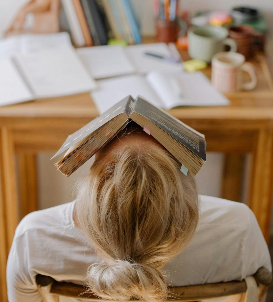 Student feeling stress and exhaustion while studying at a cluttered desk with an open book on their head.
