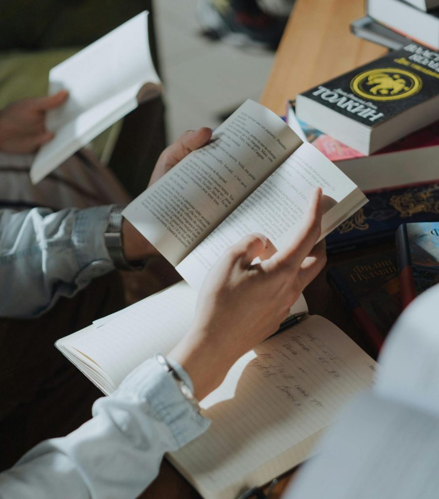 Person writing in a notebook while reading, surrounded by books in a cozy study setting.
